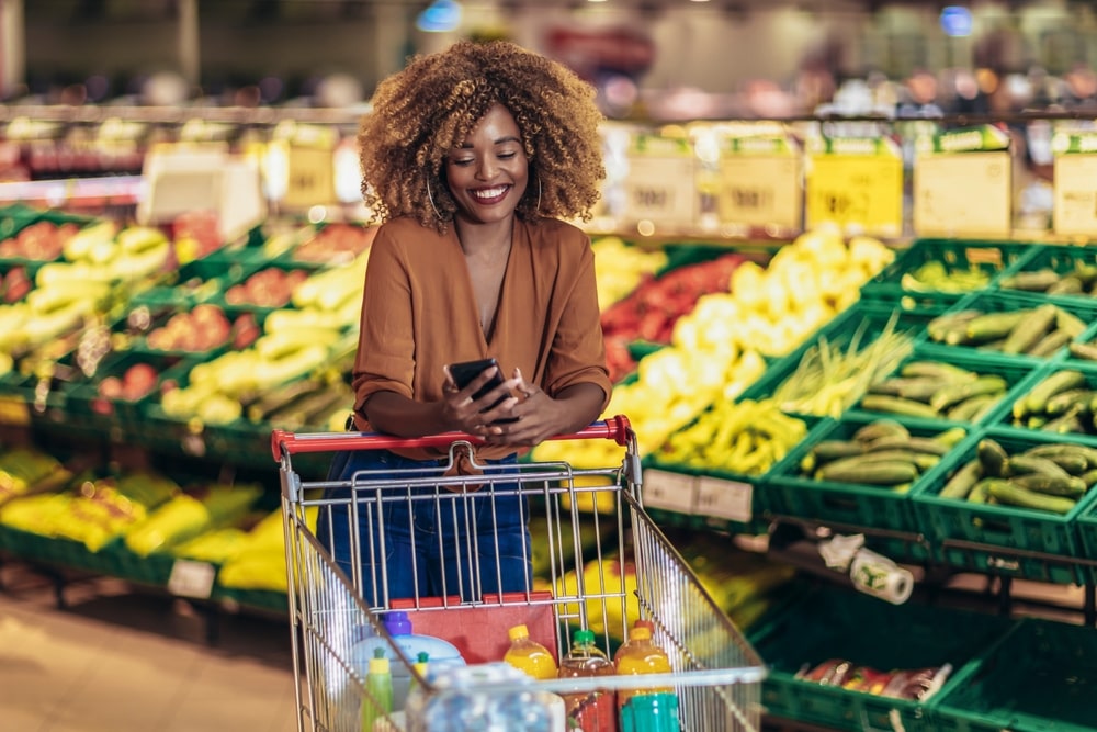 Cheerful African American Woman In Supermarket Choosing Fresh Grocery Using Phone